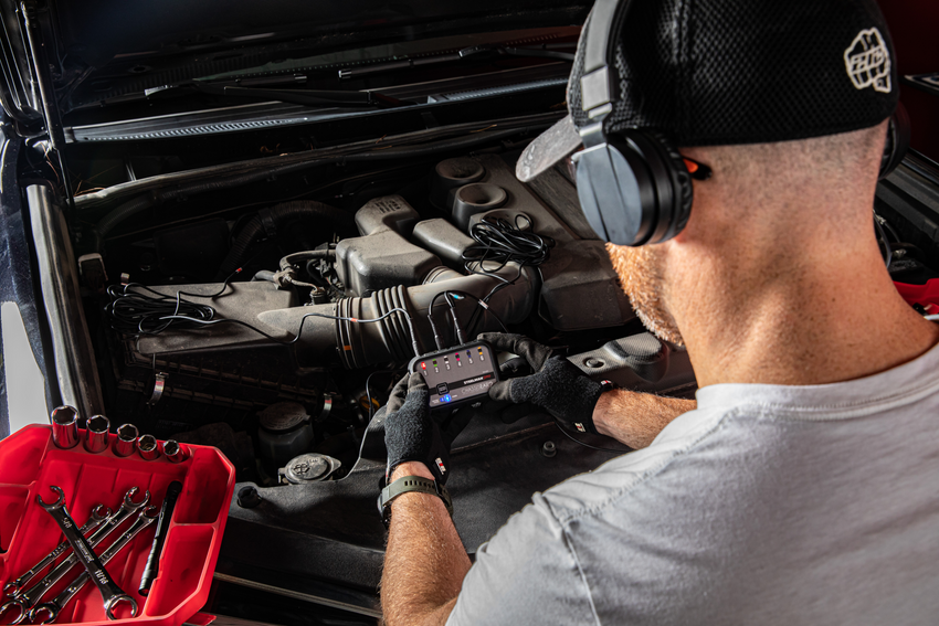 A male mechanic leans over the engine compartment of a vehicle while using the STEELMAN ChassisEAR 2 Automotive Diagnostic Noise Finder. He is wearing headphones and holding the main control unit.