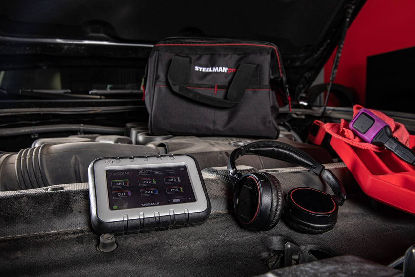 The control monitor, a transmitter, and a pair of headphones from the STEELMAN Wireless ChassisEAR 2 Automotive Diagnostic Noise Finder sit on top of a vehicle’s engine compartment.