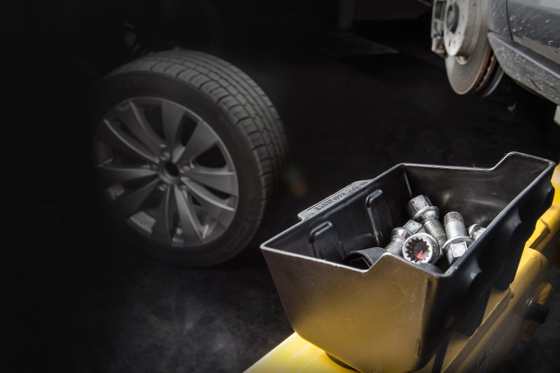 A grey STEELMAN storage bin holding lug nuts that have been removed from the vehicle wheel in the background.