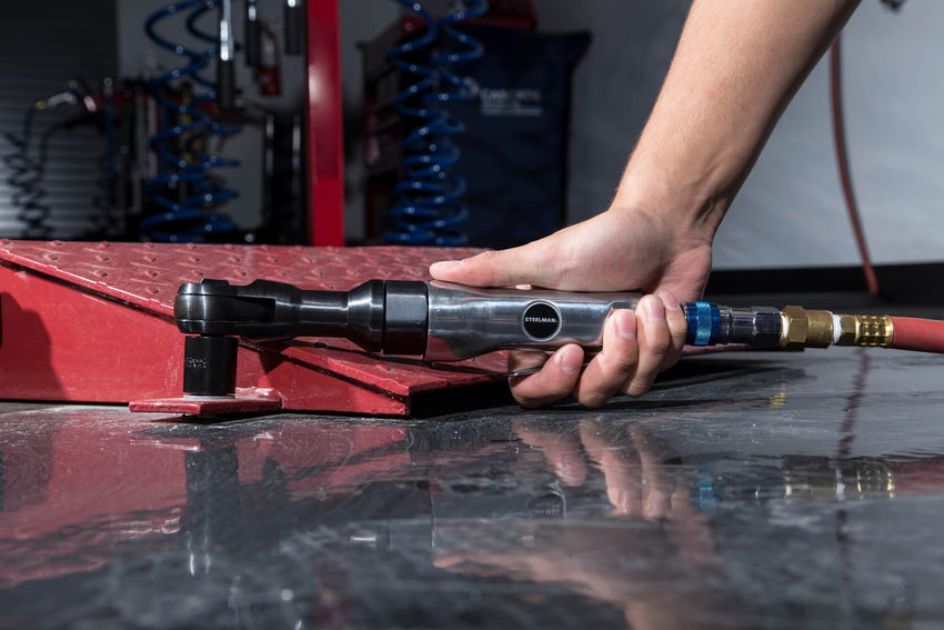 A mechanic uses a STEELMAN pneumatic ratchet to adjust a fastener on a metal ramp in an auto shop