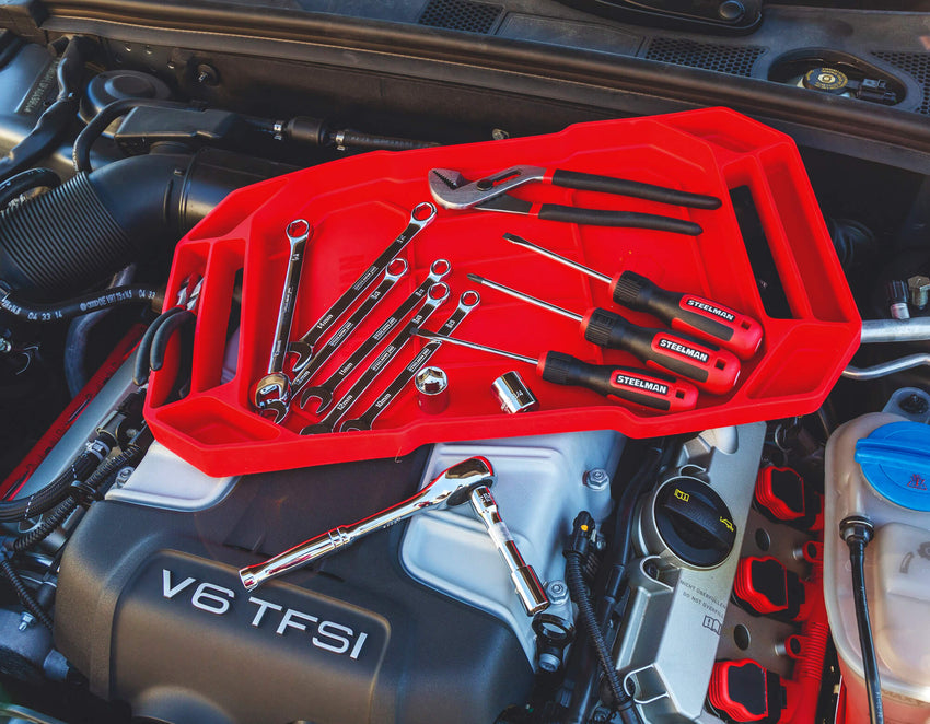 A red silicon organizer tray sits on top of a car’s engine compartment. The tray is flexible and grips the surfaces it is placed on. It’s holding wrenches, screwdrivers, pliers, and sockets.